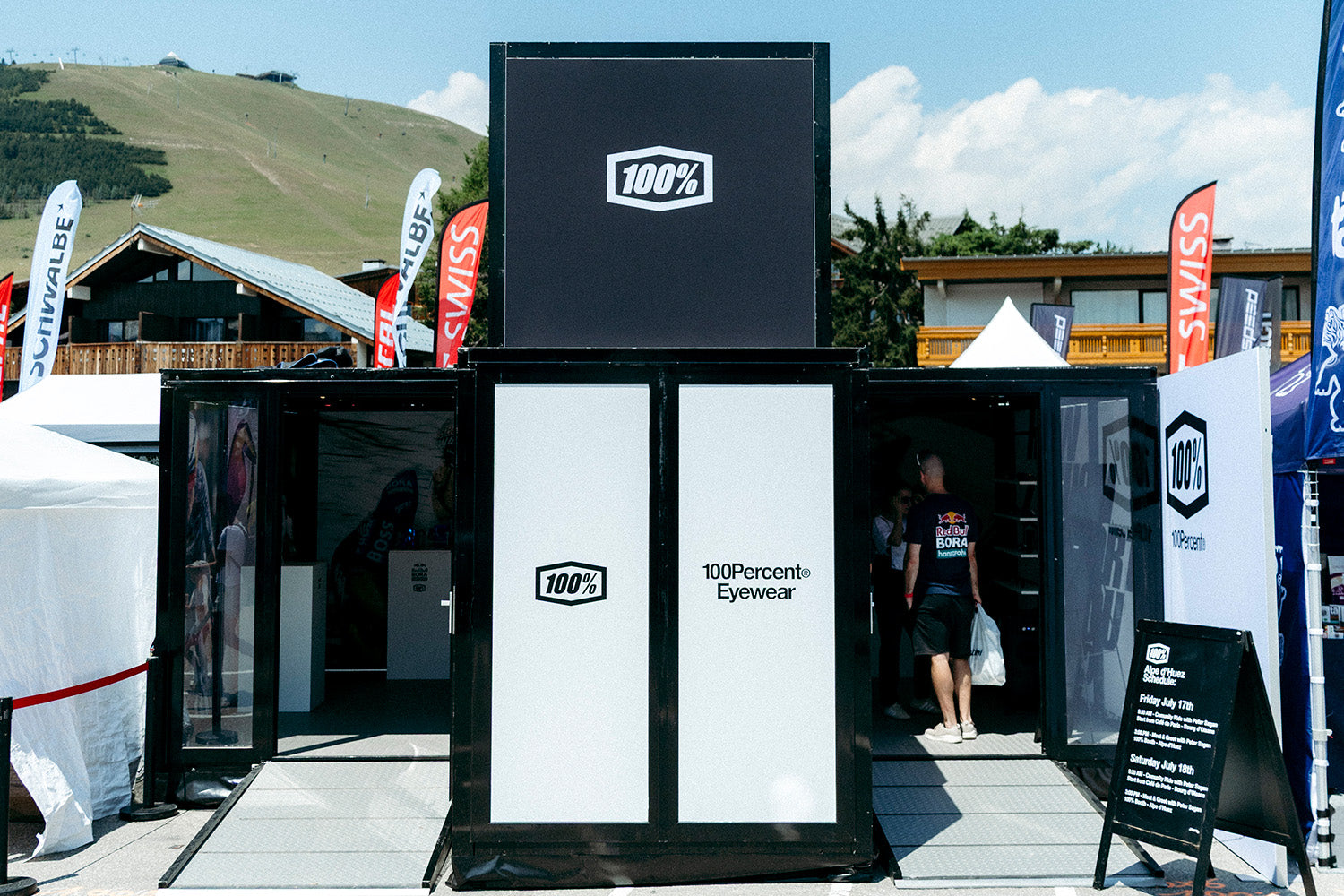The black 100Percent® Eyewear pop-up booth at Alpe d'Huez, its A-frame sign listing Peter Sagan meet-and-greets, with sponsor flags and mountains behind.