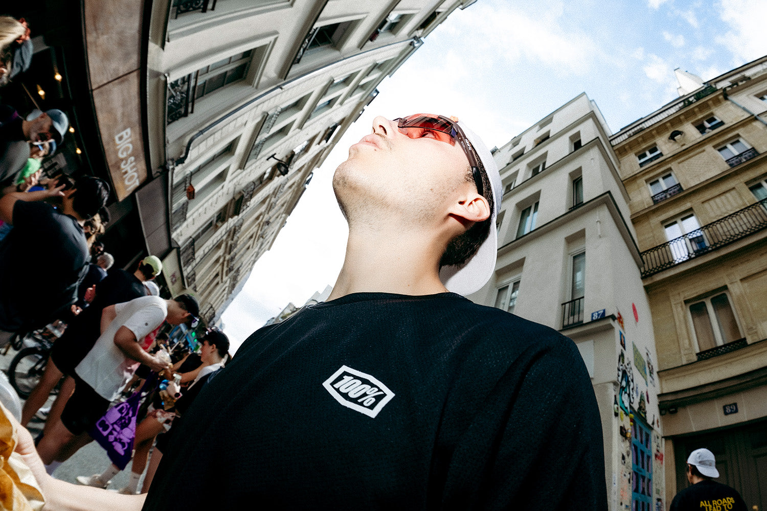 Runner in a black long-sleeve tee gazing up in red 100Percent A2 sunglasses, framed by Paris buildings.