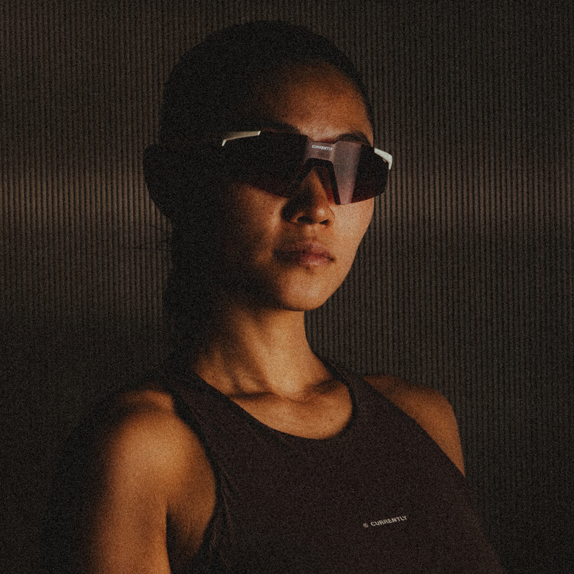 Currently Running™ x 100Percent® Collection - Featuring a girl runner wearing a Currently Running top and 100Percent A2 Sunglasses on a dark-warm moody ripple background.